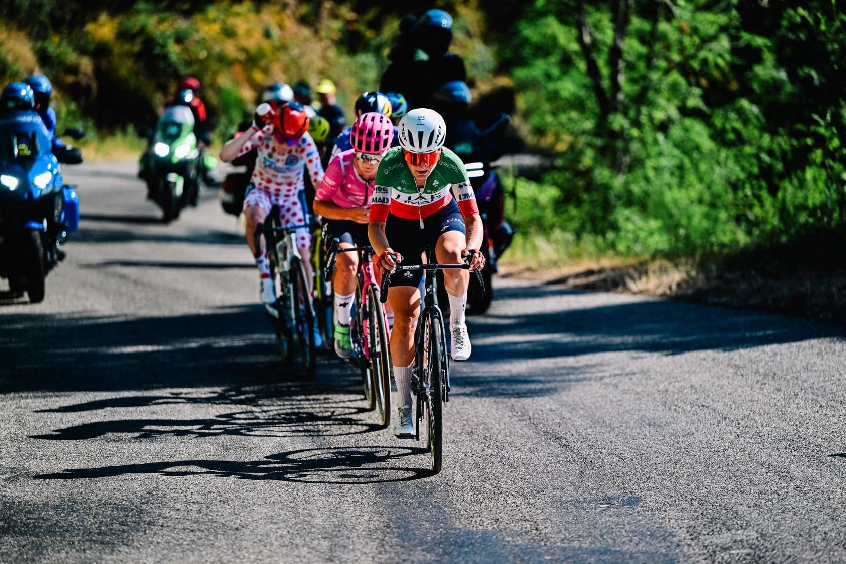 Cyclists race uphill on a road, with a rider in green-white-red jersey leading the peloton.