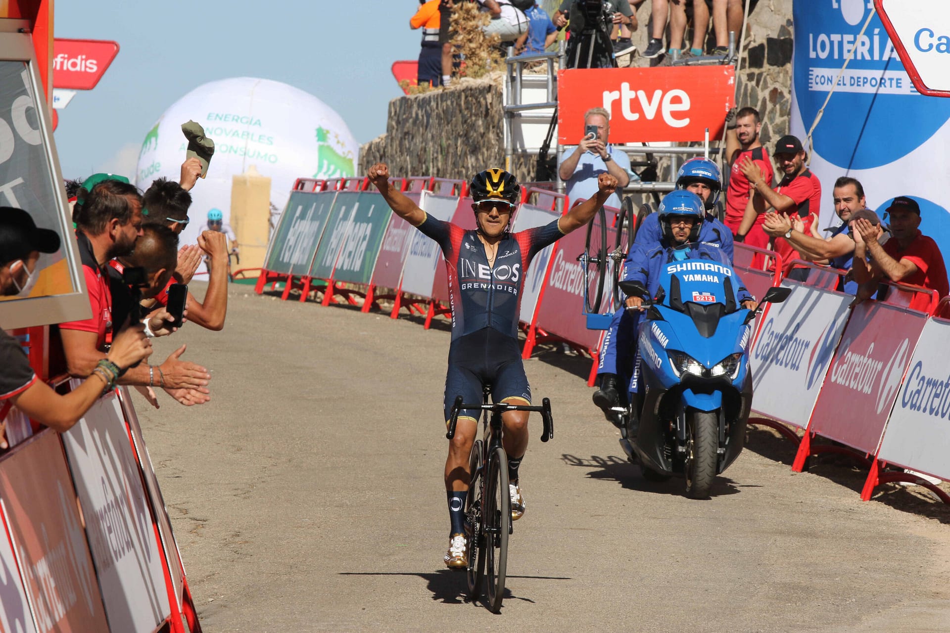 INEOS Grenadiers cyclist celebrates while riding through cheering fans beside a race motorcycle
