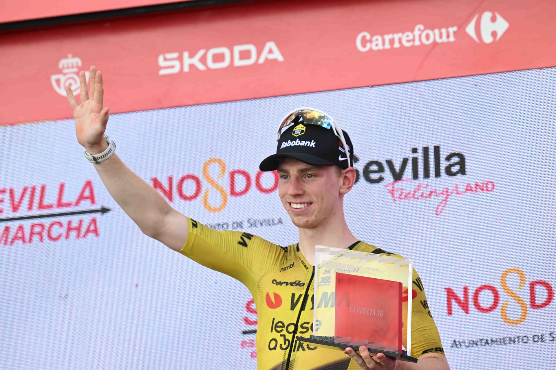 Cyclist in a yellow jersey waves while holding an award on a podium in Seville