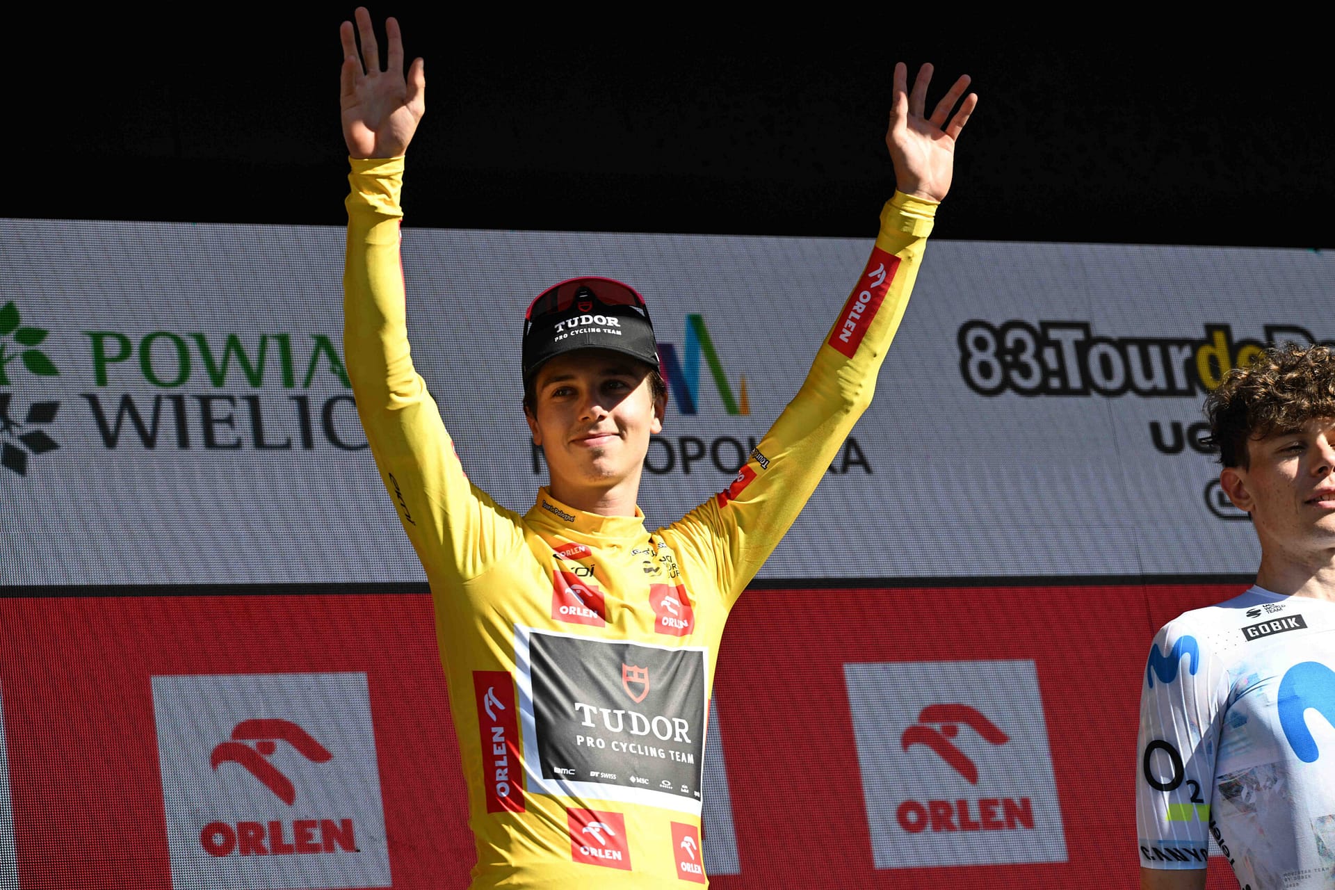 Cyclist in yellow jersey raises both arms on the Tour de France podium, smiling in celebration.