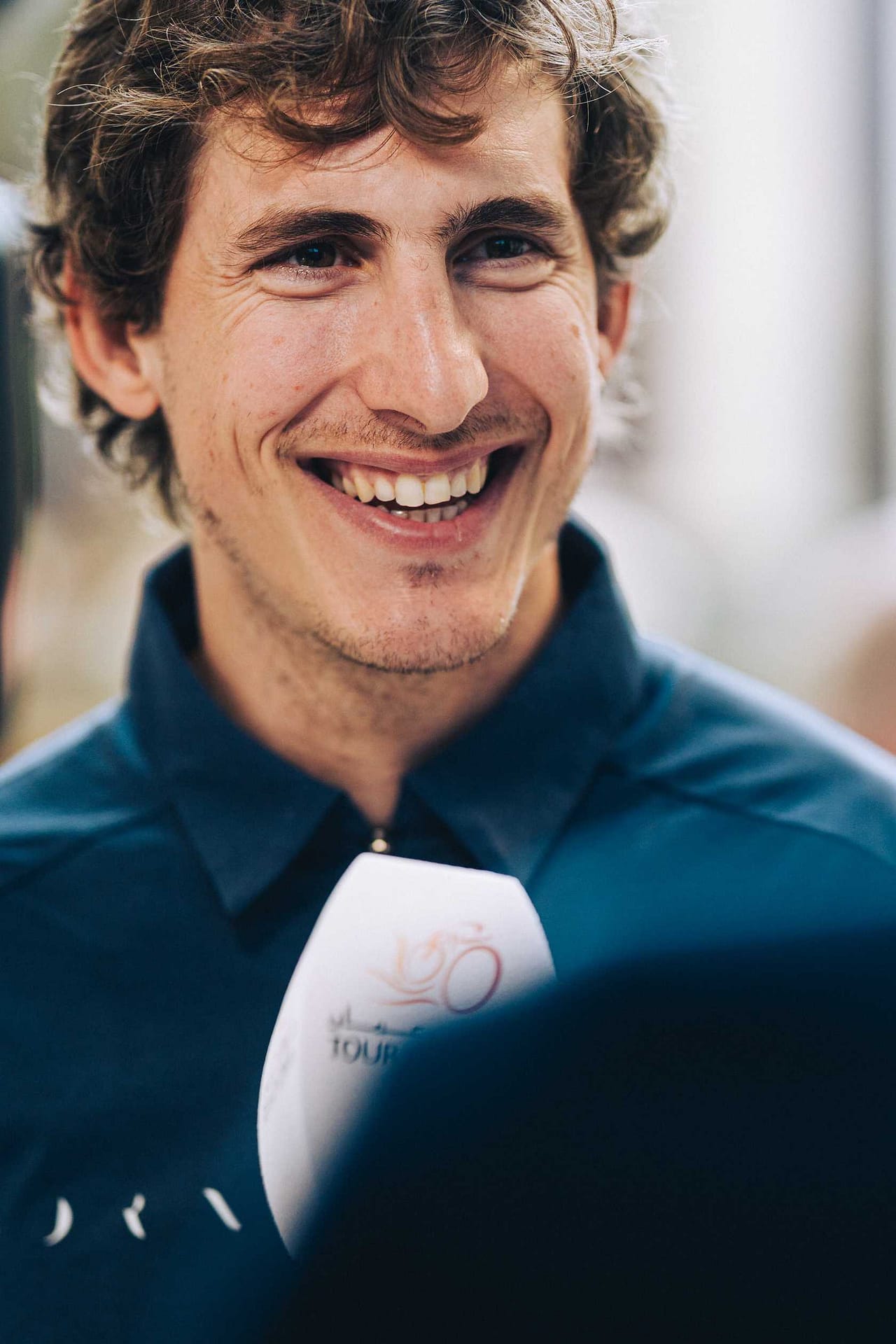 Smiling curly-haired cyclist in a blue jacket, close-up portrait with race photo in hand