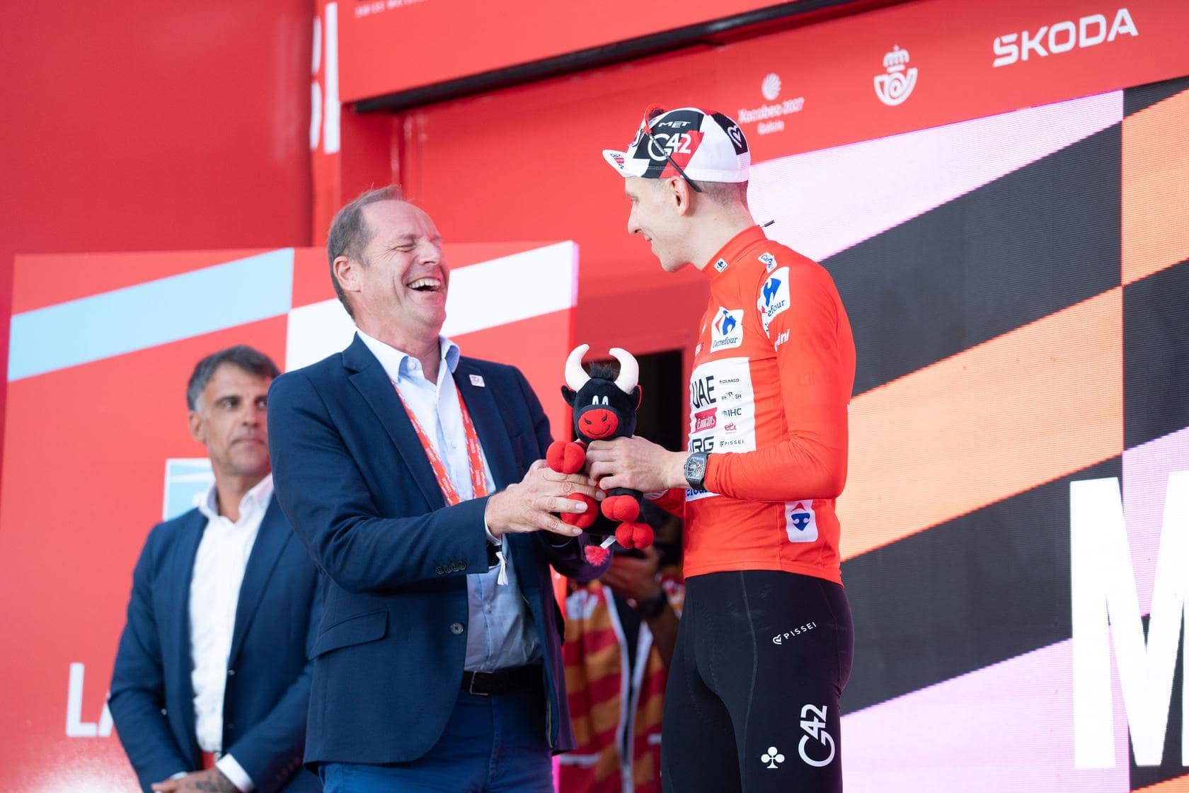 Professional cyclist in red jersey receives a plush trophy from an official on a podium.