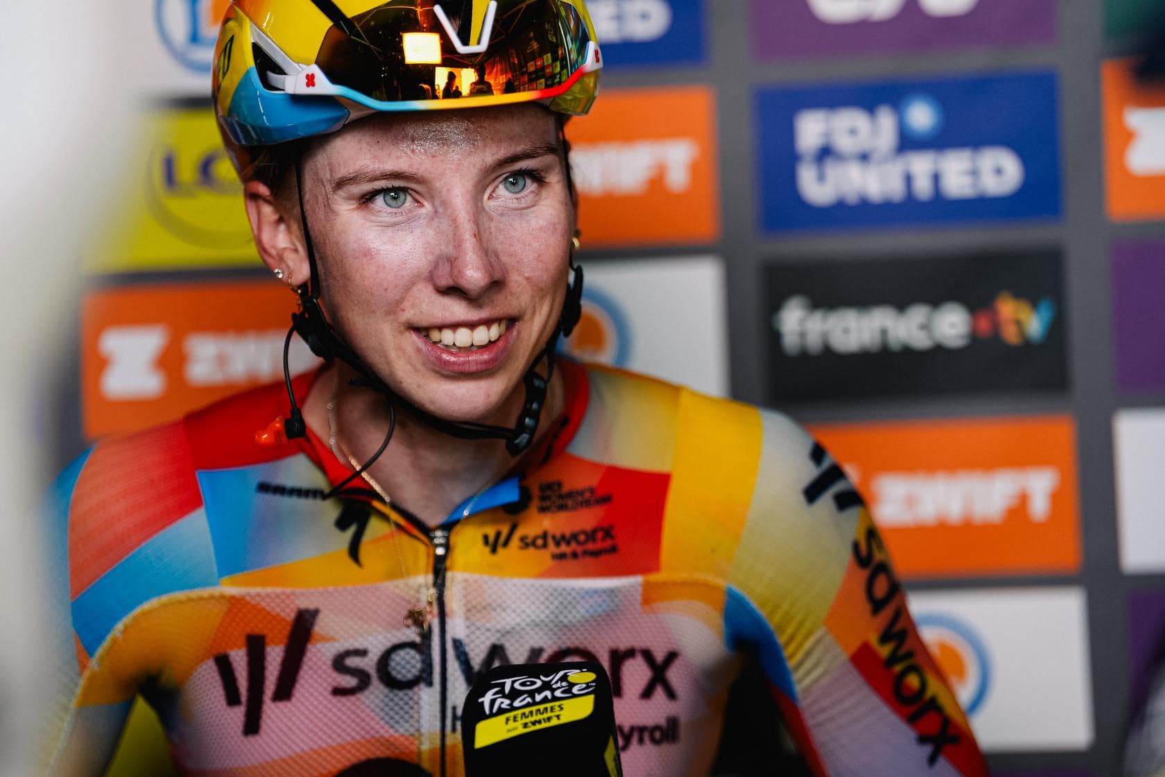 Cyclist in rainbow jersey and helmet smiles at the finish line, speaking to the camera