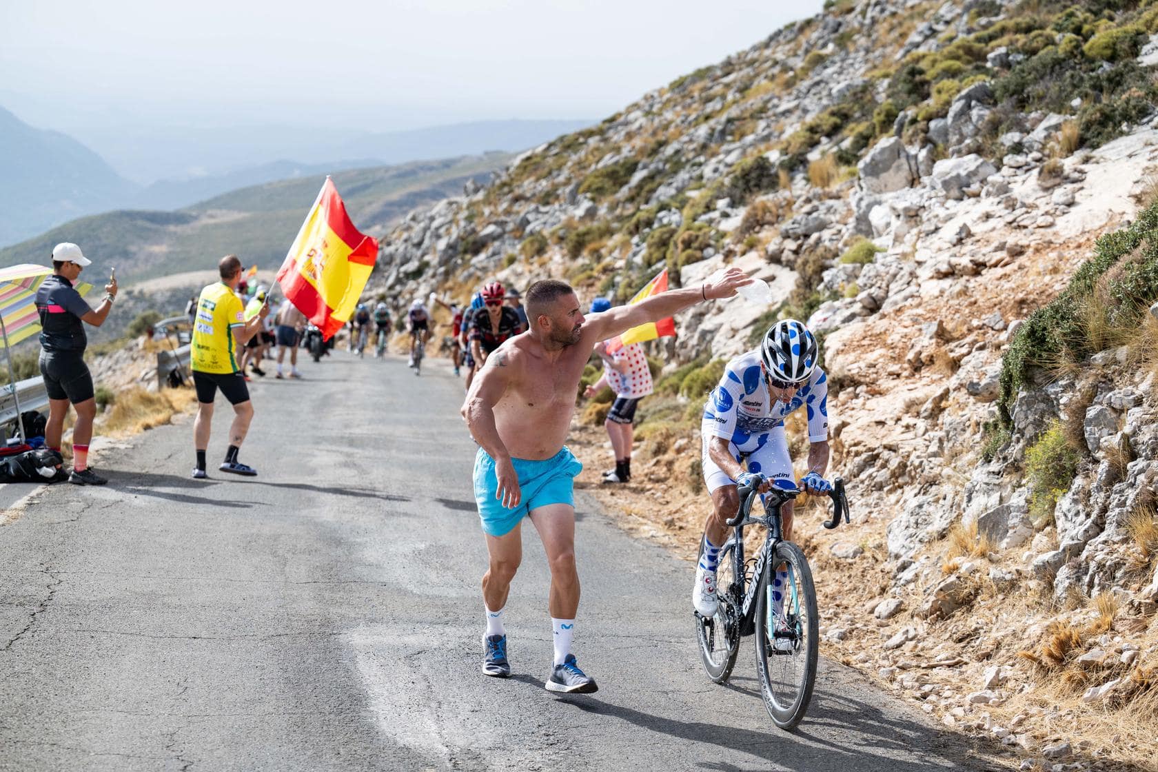 Renner zwoegt op een zonnige bergklim terwijl een toeschouwer langs de Vuelta-route aanmoedigt.