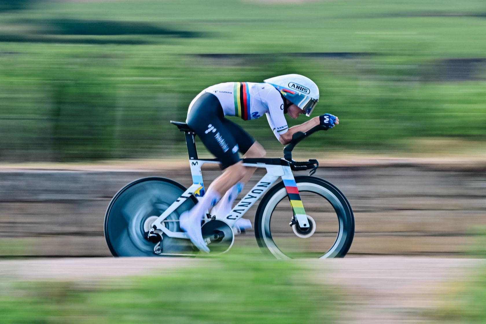 Cyclist in aero position speeds along road during a race, blurred countryside background.