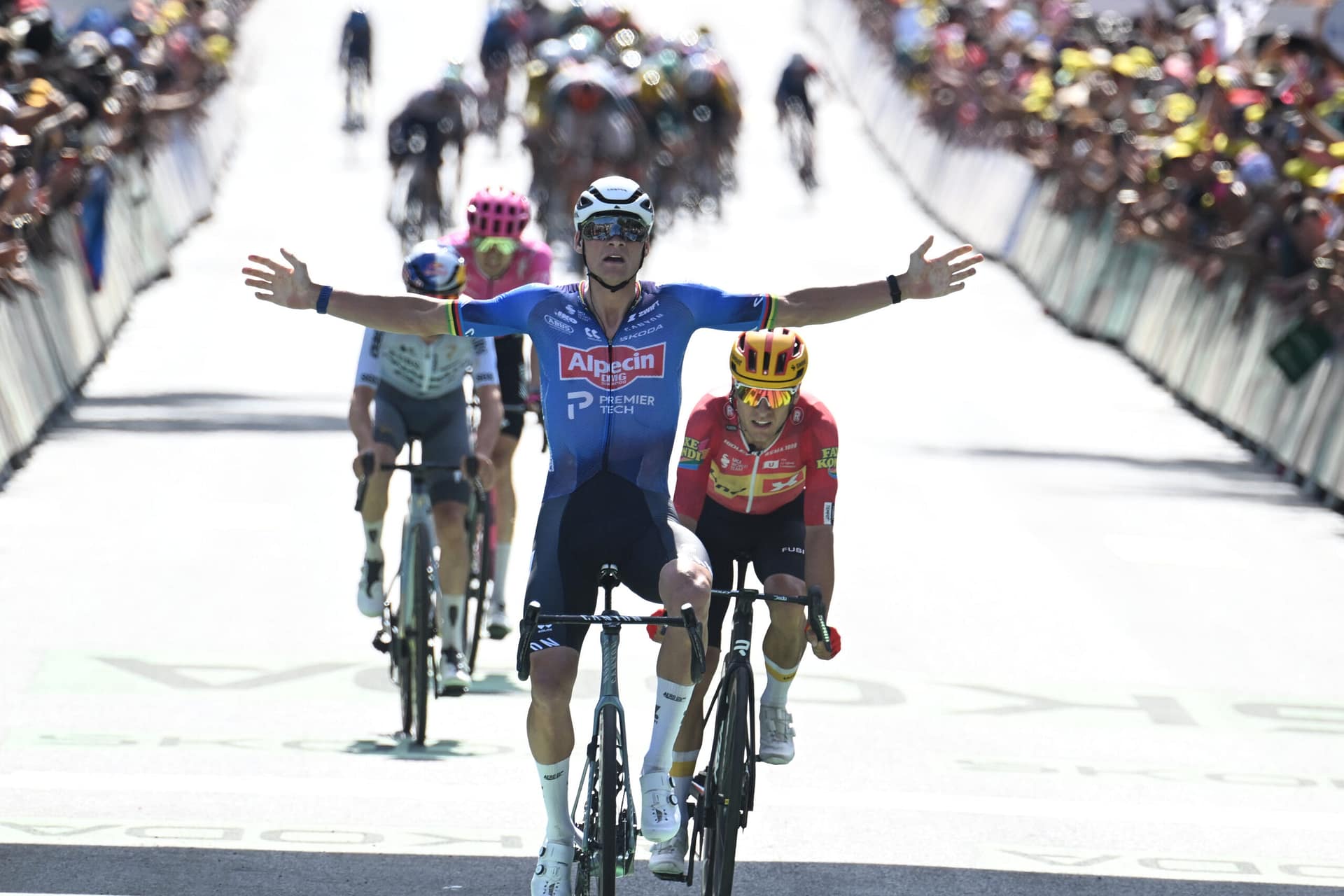 Cyclist in blue raises arms on the final stretch, leading a sprint peloton under race banners