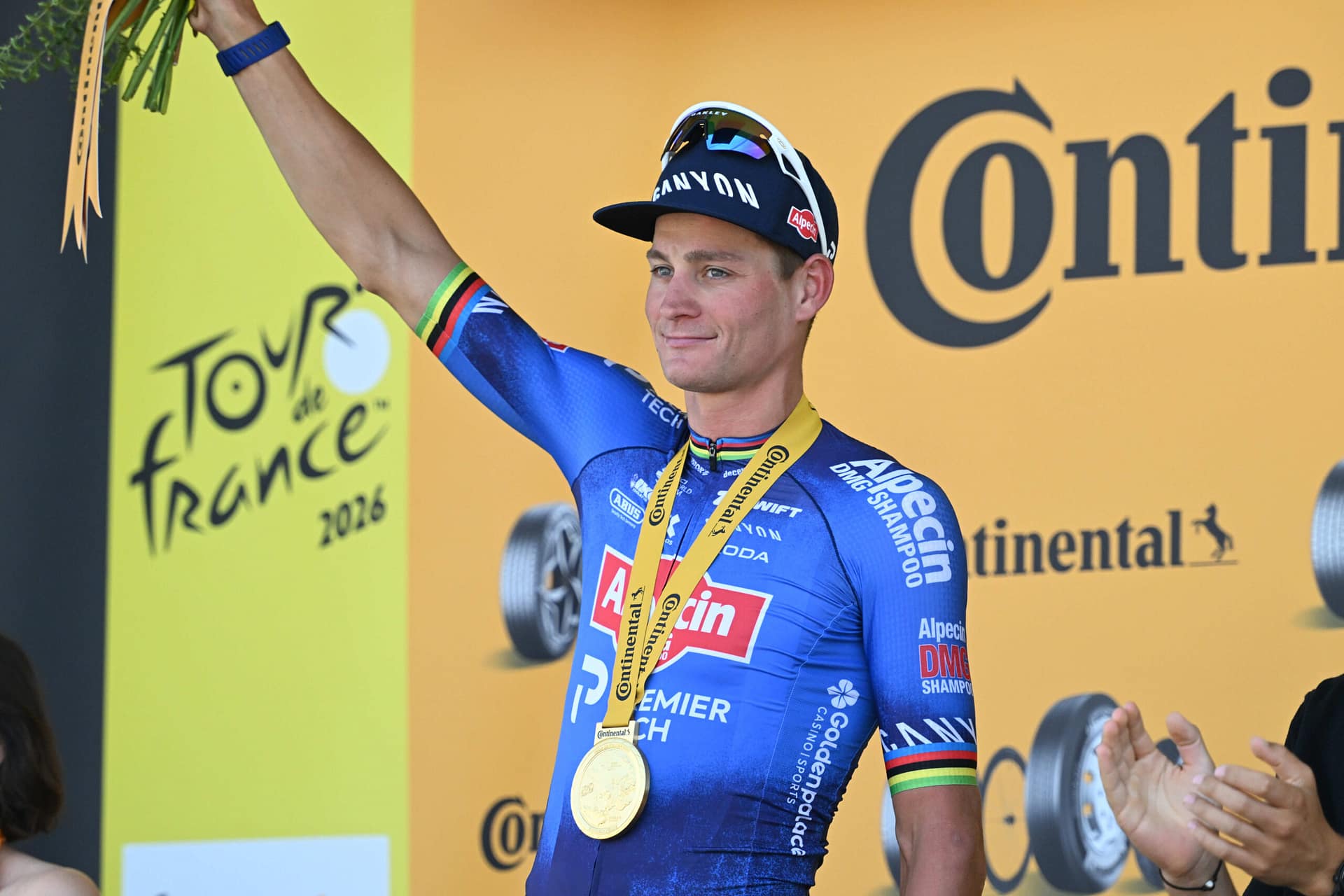 Cyclist in blue kit raises arm while holding Tour de France trophy on podium