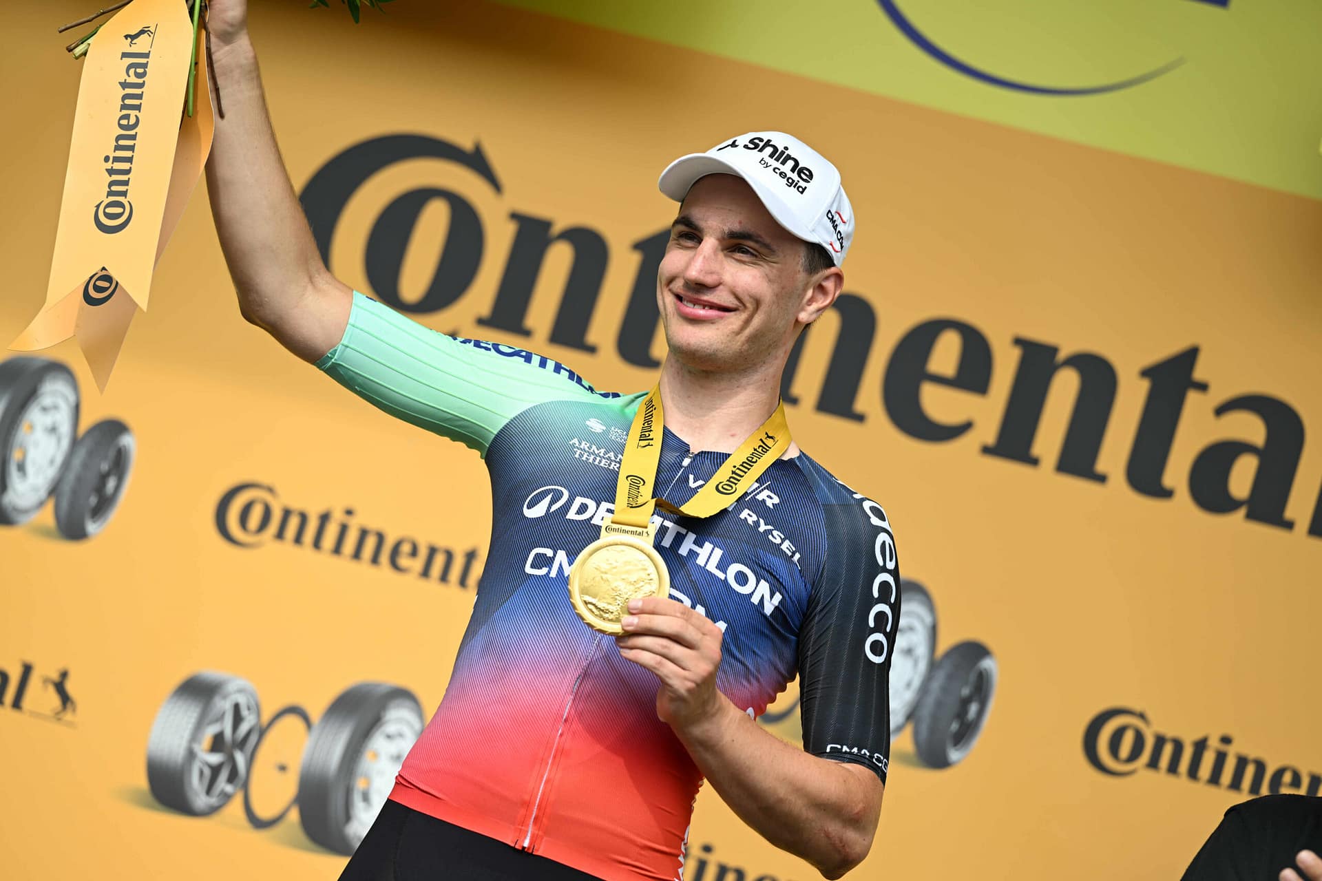 Cyclist in team kit celebrates with a gold medal on the podium, raising an arm.