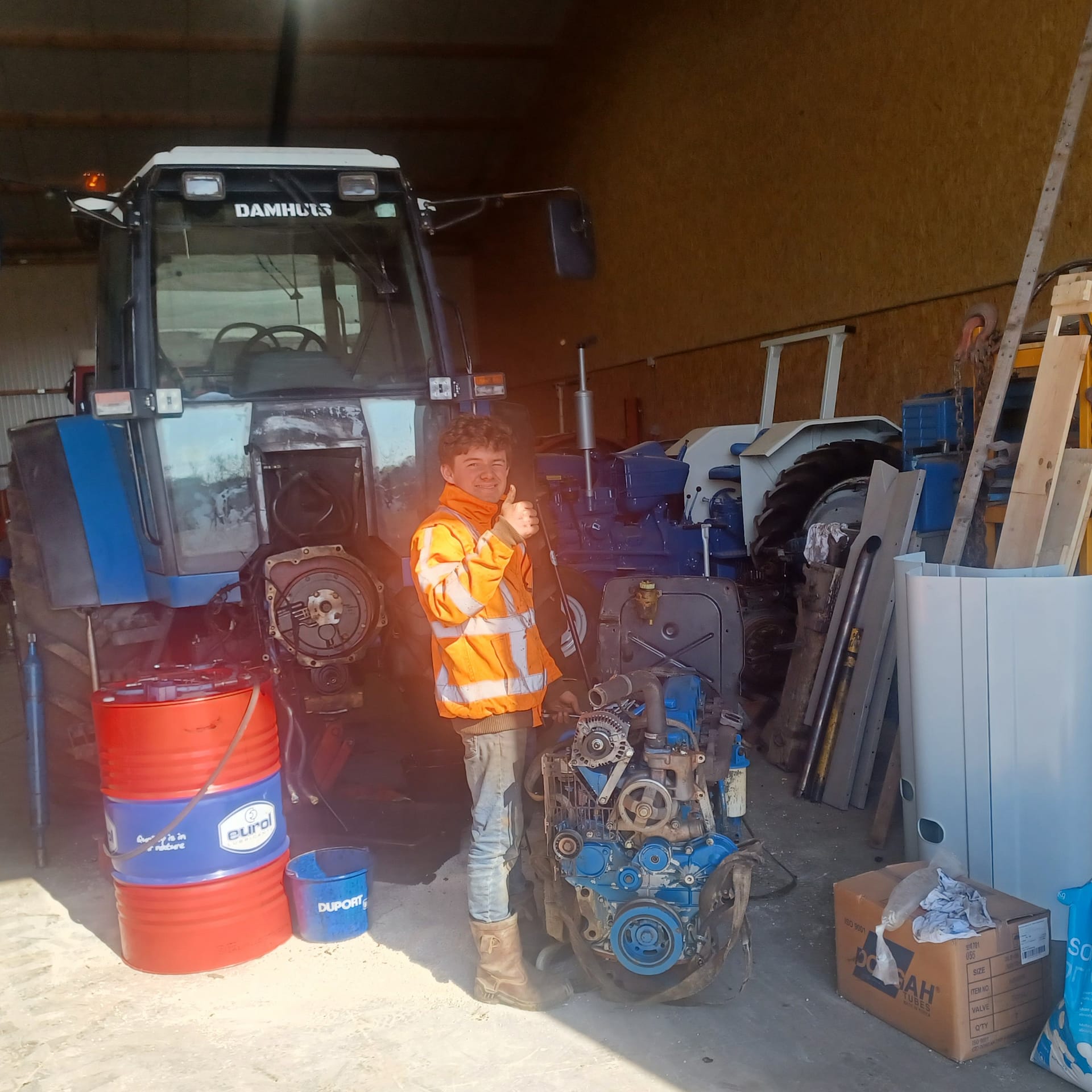 Jonge monteur in oranje werkjas sleutelt aan een tractor in een werkplaats. Op de voorgrond staat een motorblok en vaten met olie.