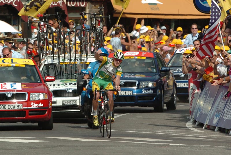 Cyclist in yellow rides past team cars at the Tour de France stage finish in Morzine, cheered by fans.