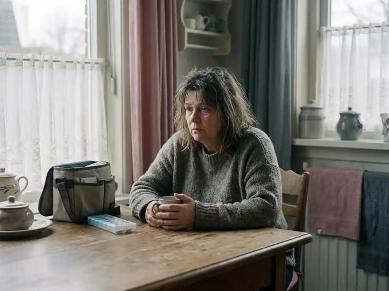 Uitgeputte vrouw aan keukentafel met onaangeroerde thee, mantelzorgtas en medicijndoos zichtbaar, zacht middaglicht door kanten gordijnen.