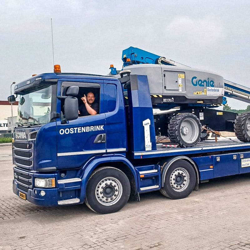 vrachtwagenchauffeur werkzaam in de logistiek zit in blauwe vrachtwagen en steekt hand op om te begroeten.