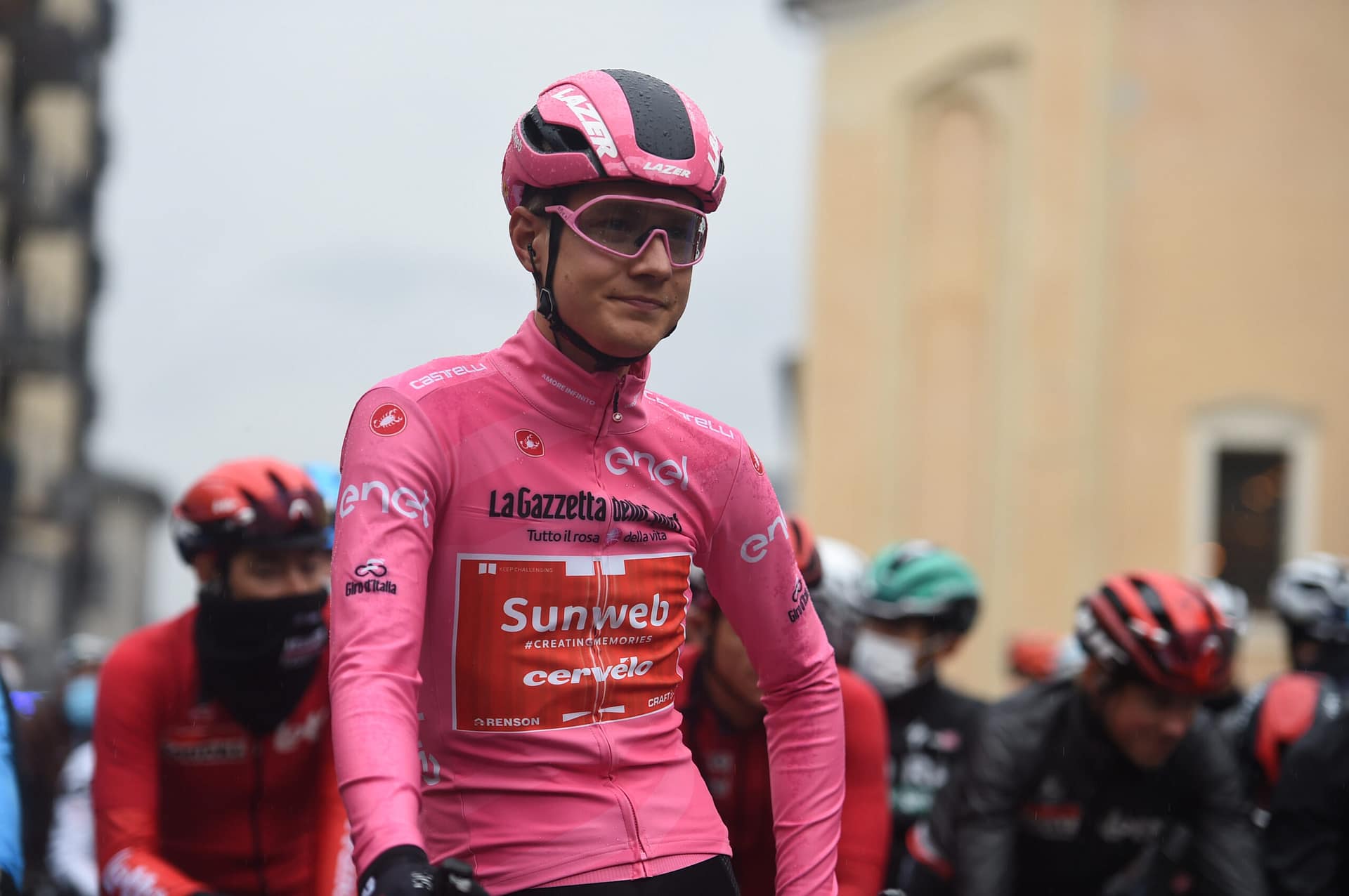 Cyclist in pink team kit stands on street during race, with blurred competitors nearby