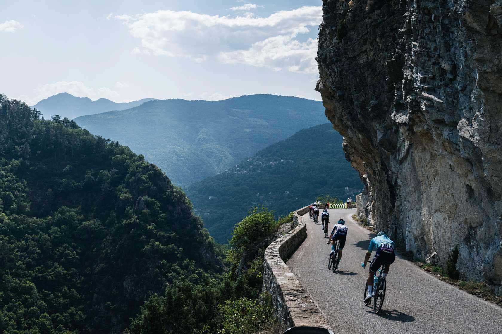 Cyclists ride along a narrow mountain road beside a cliff during a scenic Tour-style stage