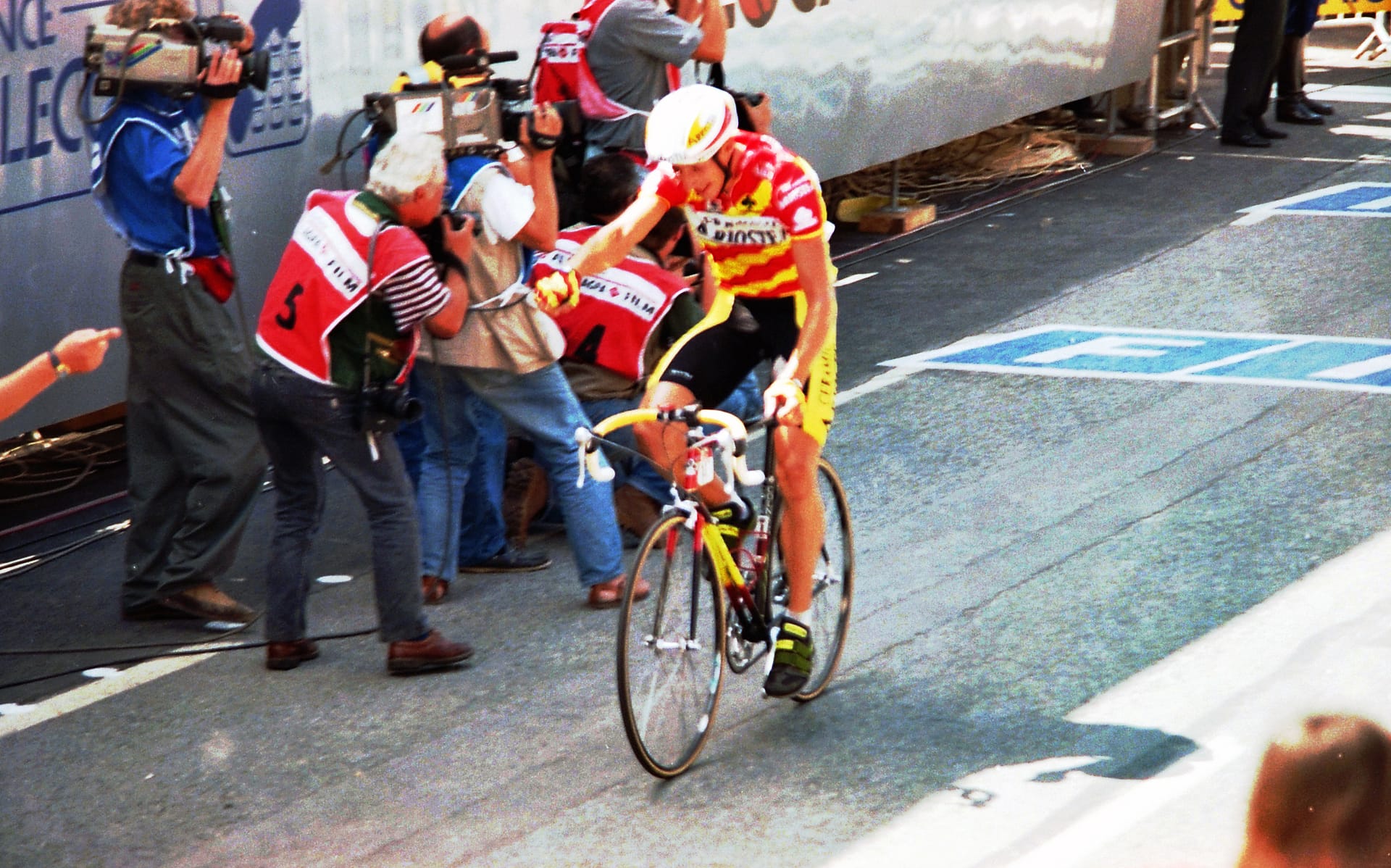 Cyclist in yellow and red reaches the finish as team staff and riders gather at Tour de France stage